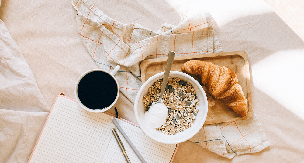 Overhead view of a notebook and a pen, surrounded by a cup of coffee, a bowl of granola and a croissant on a small tray.