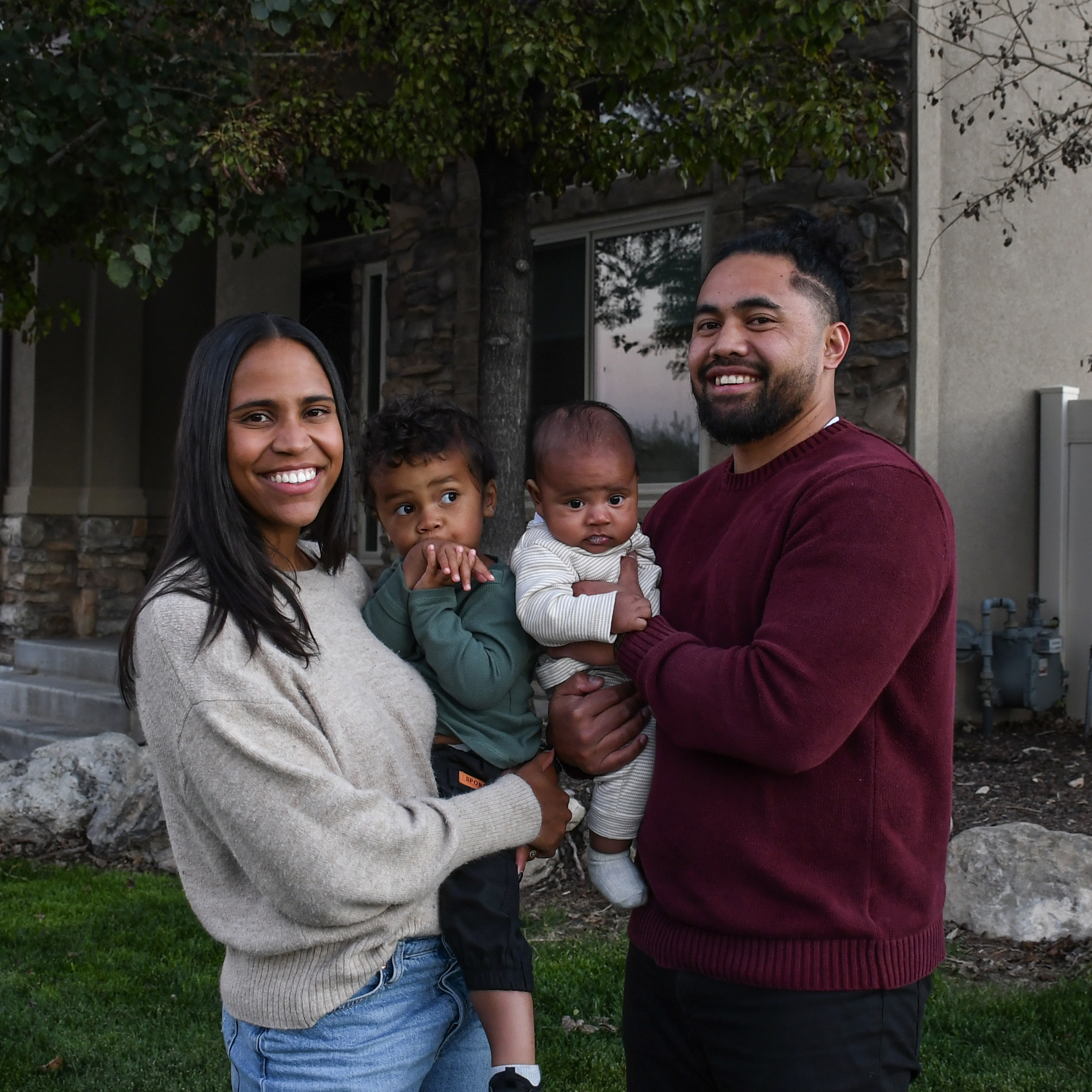 A smiling family of four standing in front of their home, with two young children, one held by the mother and the other held by the father. They are outdoors, with trees and landscaping visible.