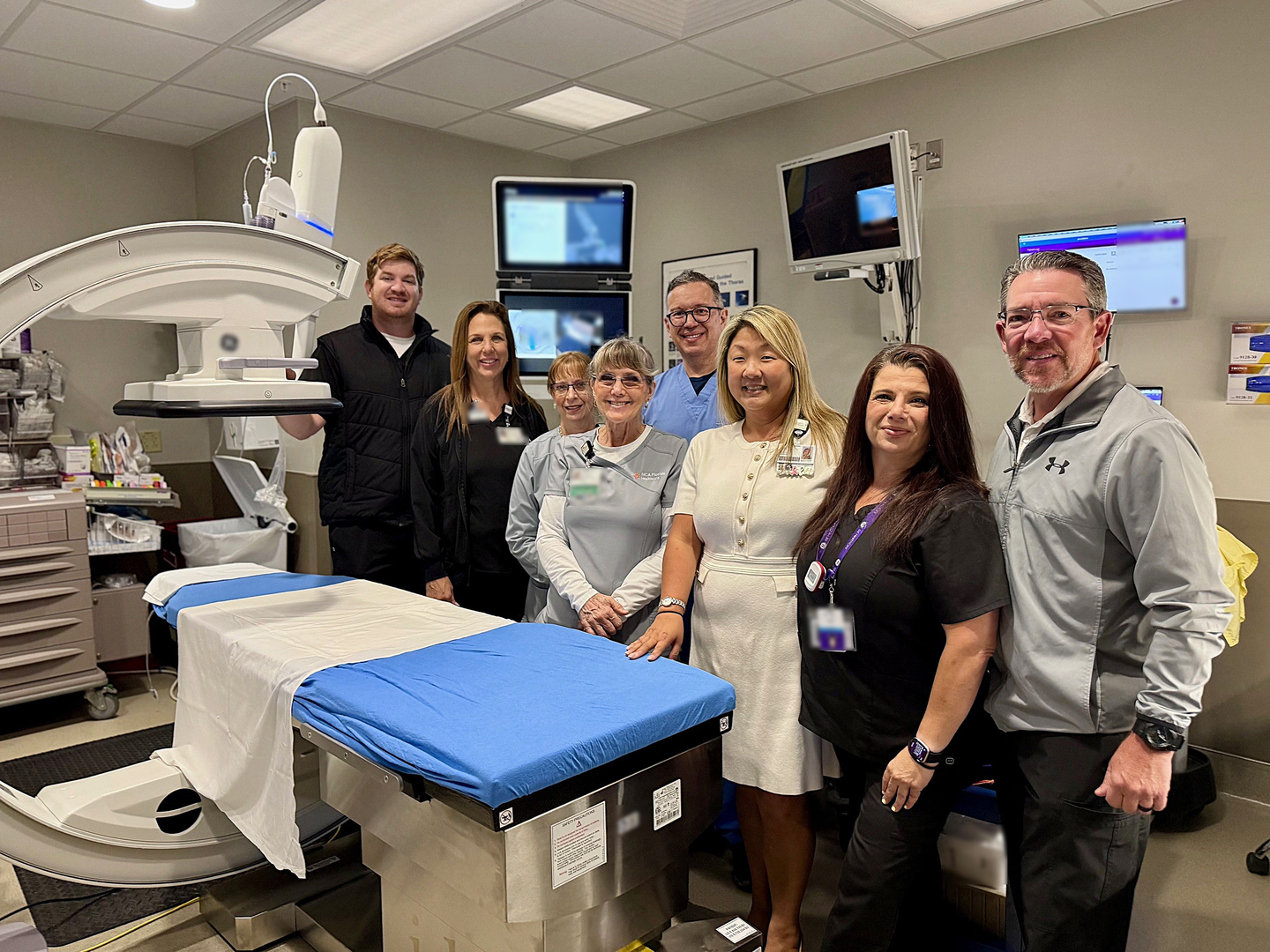 A group of healthcare staff stand together smiling in a medical procedure room beside a hospital bed and imaging equipment.
