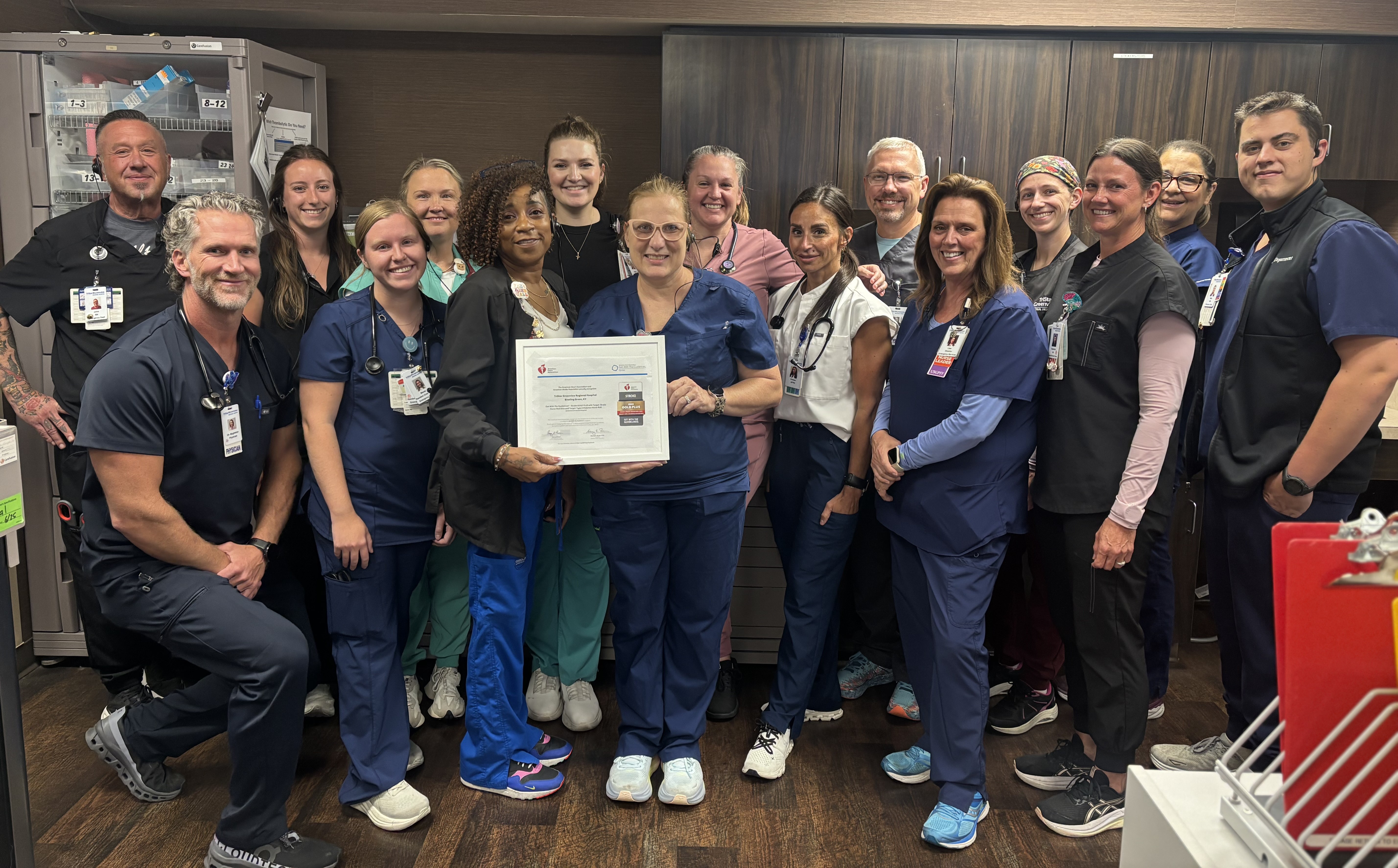 A group of approximately 16 hospital staff members in scrubs and clinical attire gather in a staff workroom to celebrate a recognition award. Two team members at the center hold a framed certificate while colleagues smile around them. Cabinets and medical supply storage are visible in the background, highlighting the clinical setting.