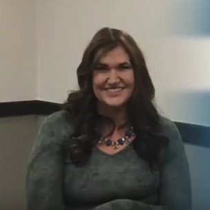 Jennifer smiles while wearing a tie dye grey shirt and a chunky gemstone necklace.