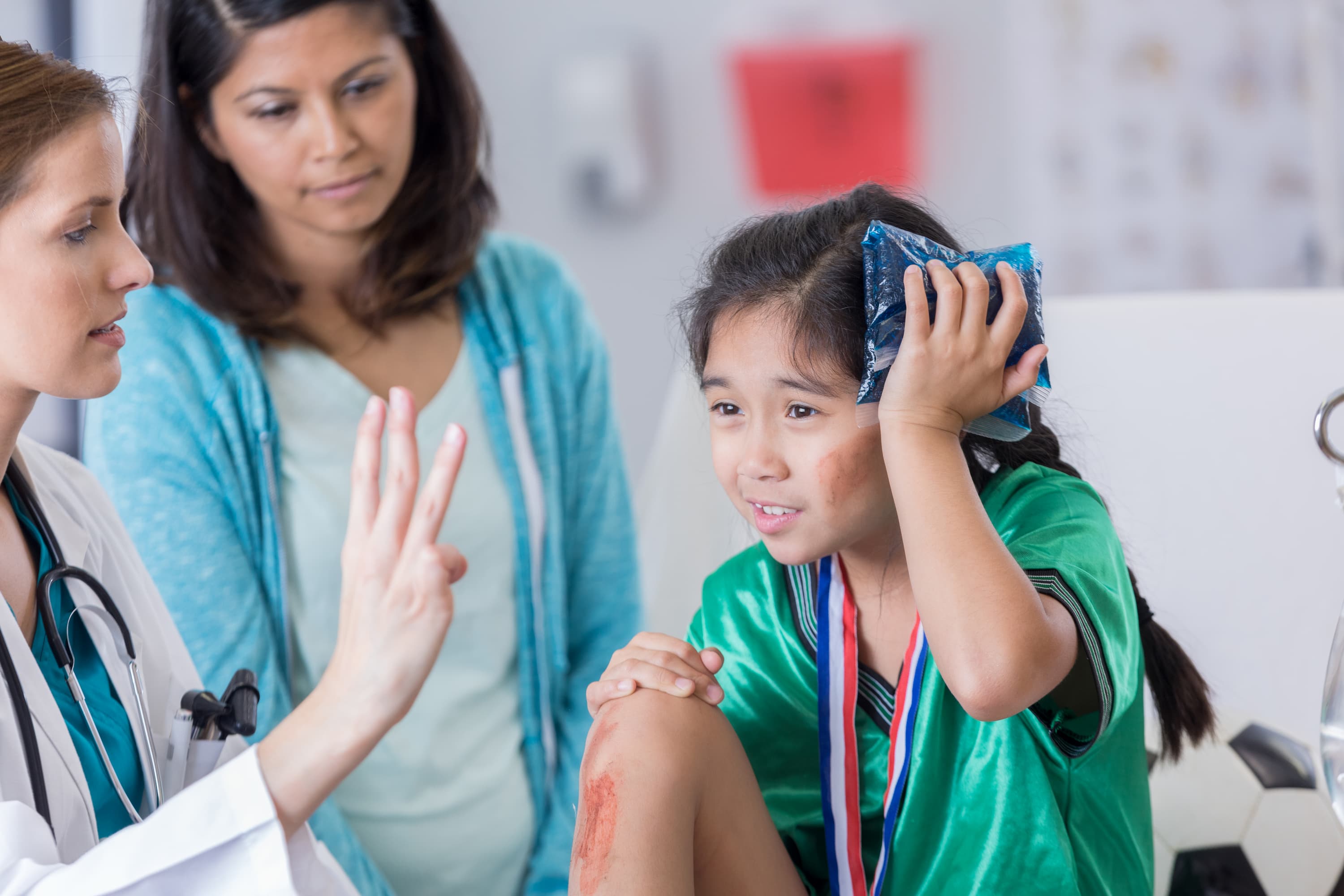 Healthcare provider examining a child with a scraped knee while the child holds an ice pack to their head, with a parent standing nearby.