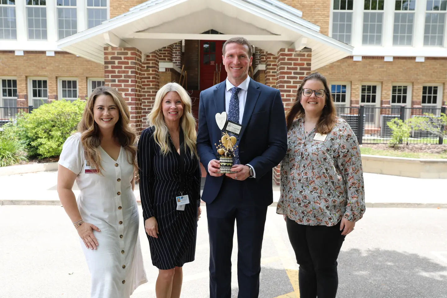 Three women and a man are standing side by side, smiling. The man is holding a black and gold award made of crayons adored with hearts.