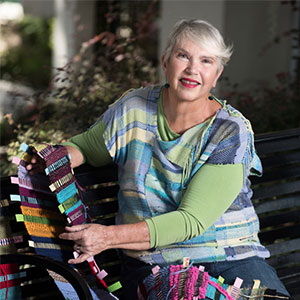 Suzanne Hokanson smiles while holding up some of her yarn crafts that she made on her loom.