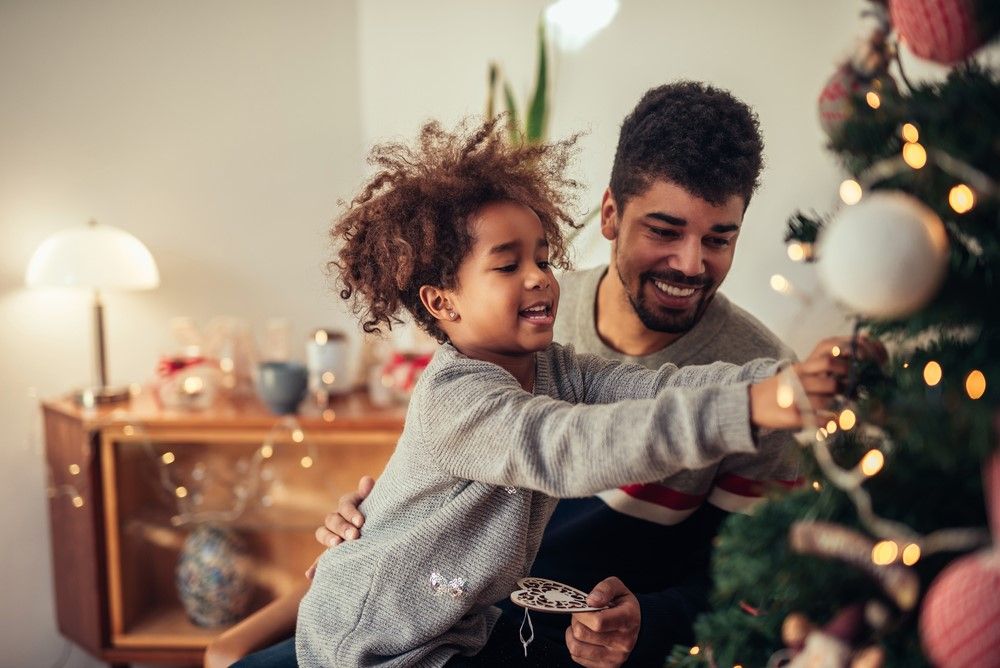A young child hangs ornaments onto a Christmas tree with their father.