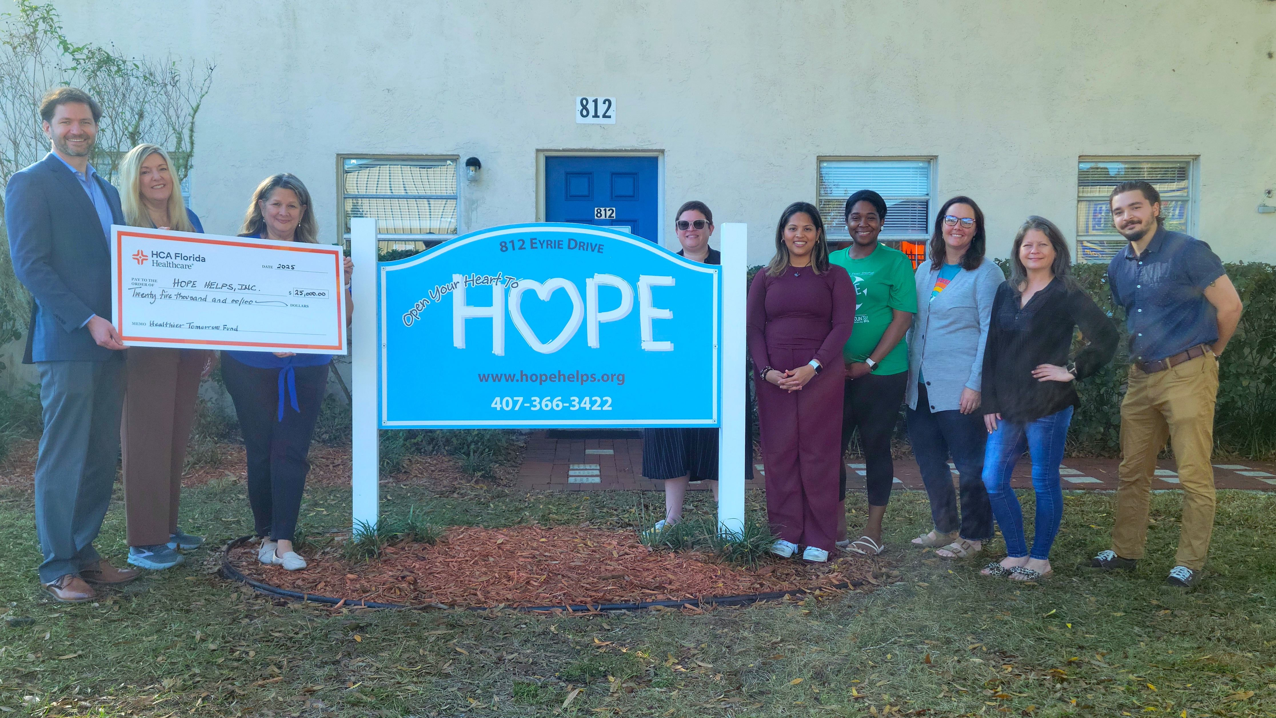 Oviedo Medical Center CEO Cameron Howard, Oviedo Medical Center Communications and Community Engagement Director Cathy Perry and HOPE Helps CEO Joan Faulkner hold a ceremonial check celebrating a $25,000 grant to help provide nutritious meals to community members in need. They are standing in front of the HOPE Helps building with a group of smiling people.