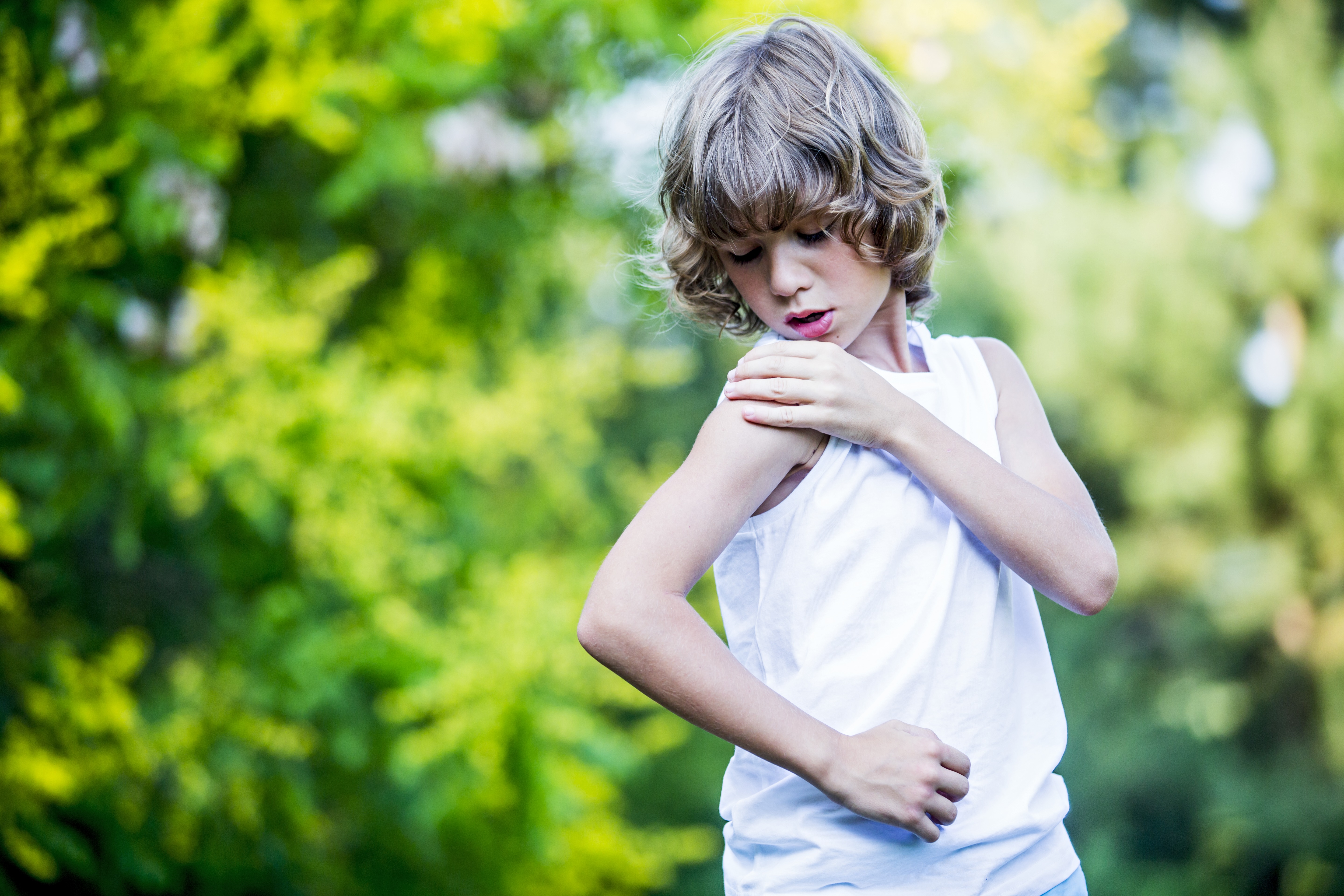 Child outdoors in a green park, looking down and touching their upper arm as if reacting to an itch or bug bite, wearing a white sleeveless shirt.