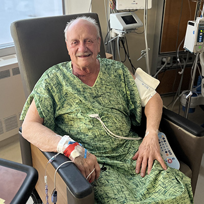Larry Buchut is smiling as he sits in a chair in a hospital room with post-surgery monitoring equipment attached to him.