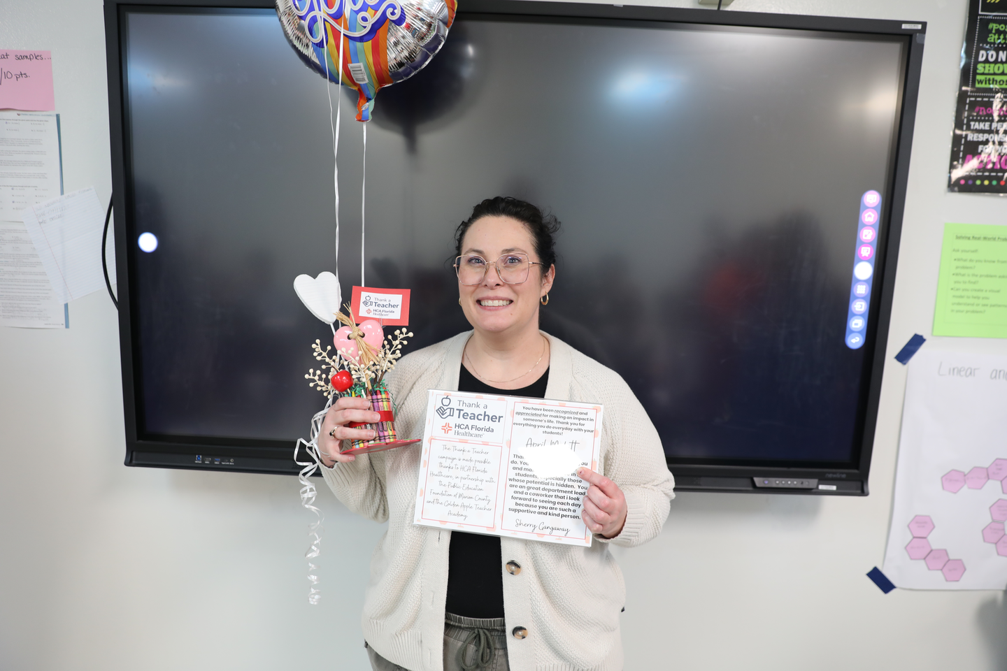 A teacher stands in front of a classroom smart board holding her "Thank a Teacher" certificate and a small decorative gift. A colorful "Thank You" balloon floats above her as she smiles for the photo during the recognition presentation.