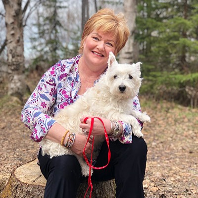 Shelly Smith is enjoying the outdoors, sitting on a tree stump and holding her dog in her lap.