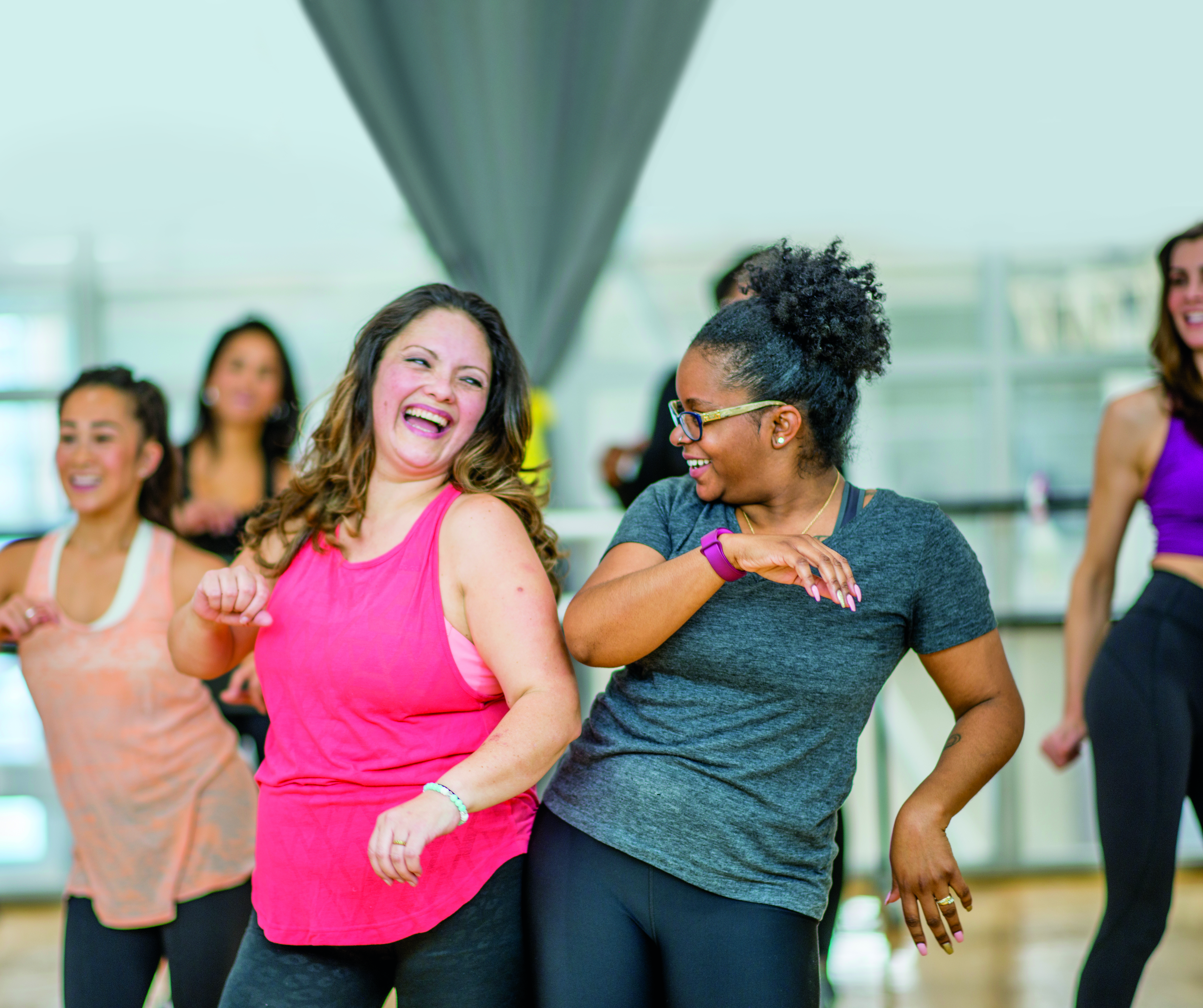A group of smiling, laughing women wearing athletic clothes are dancing in a fitness studio.