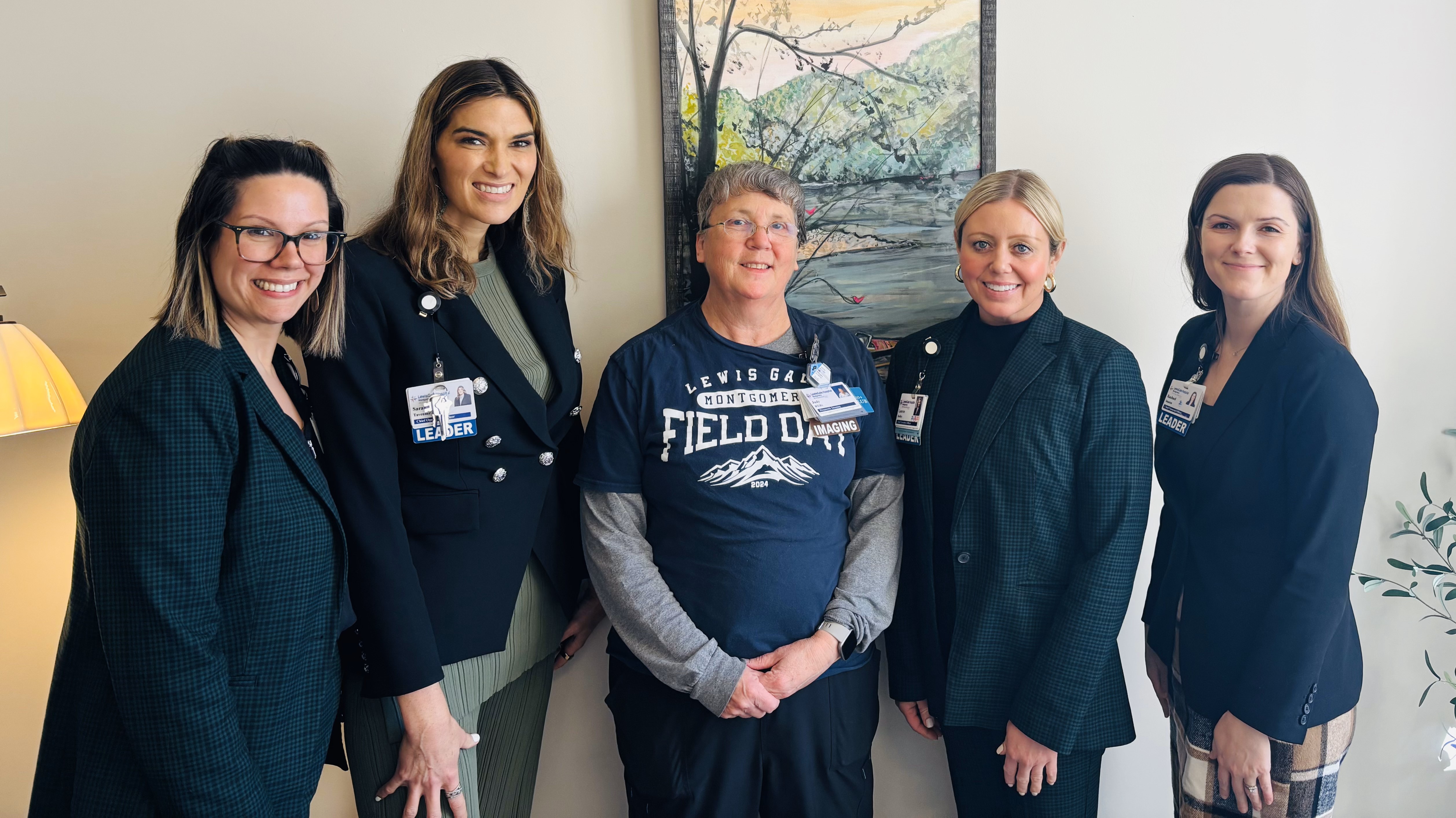 Five women standing in a row. Four are wearing professional suits and one in middle is wearing business casual clothing. They are standing in front of a framed photo of a tree.