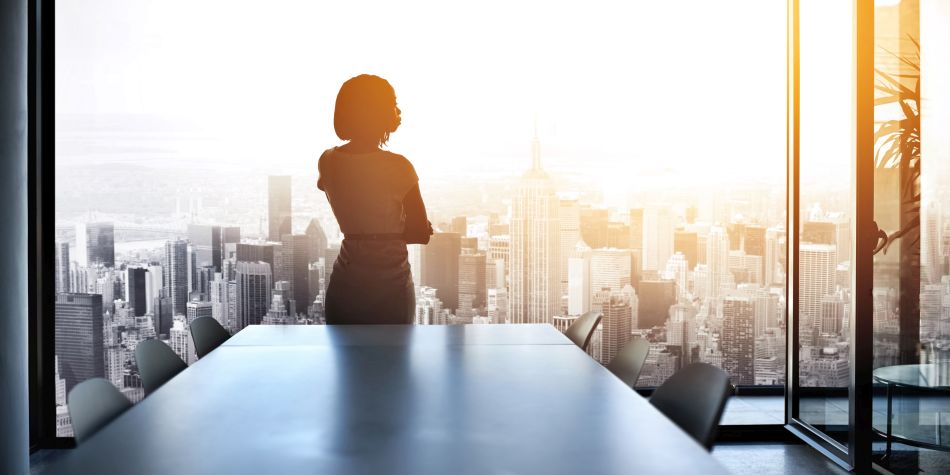 Rearview shot of a young businesswoman looking out from a large window in an office, with the cityscape in the background.