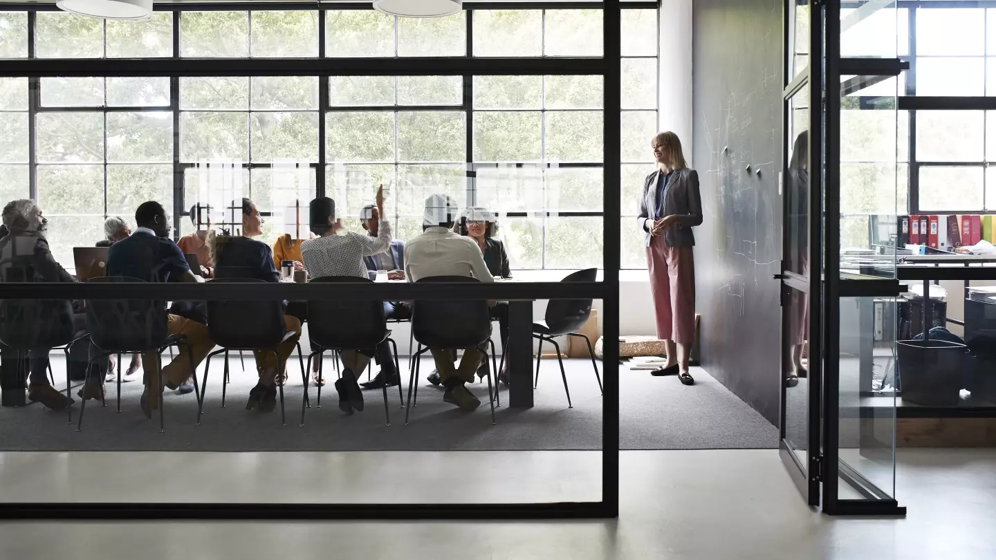 From outside an open doorway, a businesswoman presents to her colleagues in a workplace meeting.