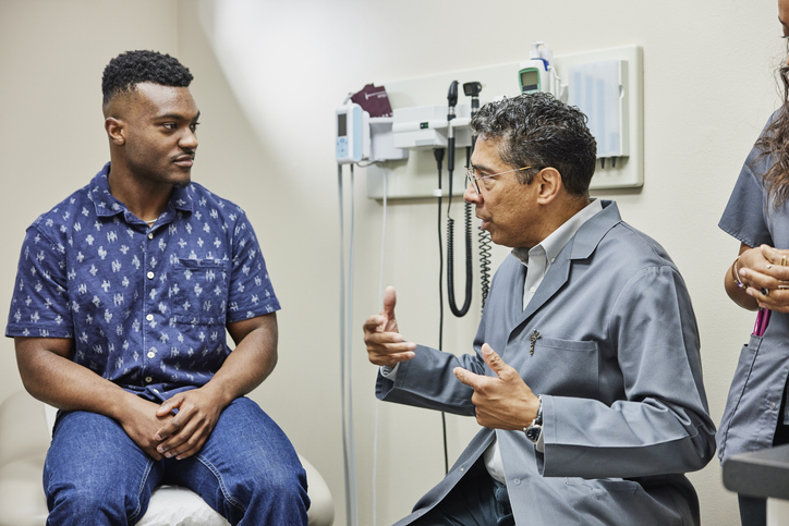 Male doctor in glasses speaking with adult male in exam room, with female healthcare professional in the room.
