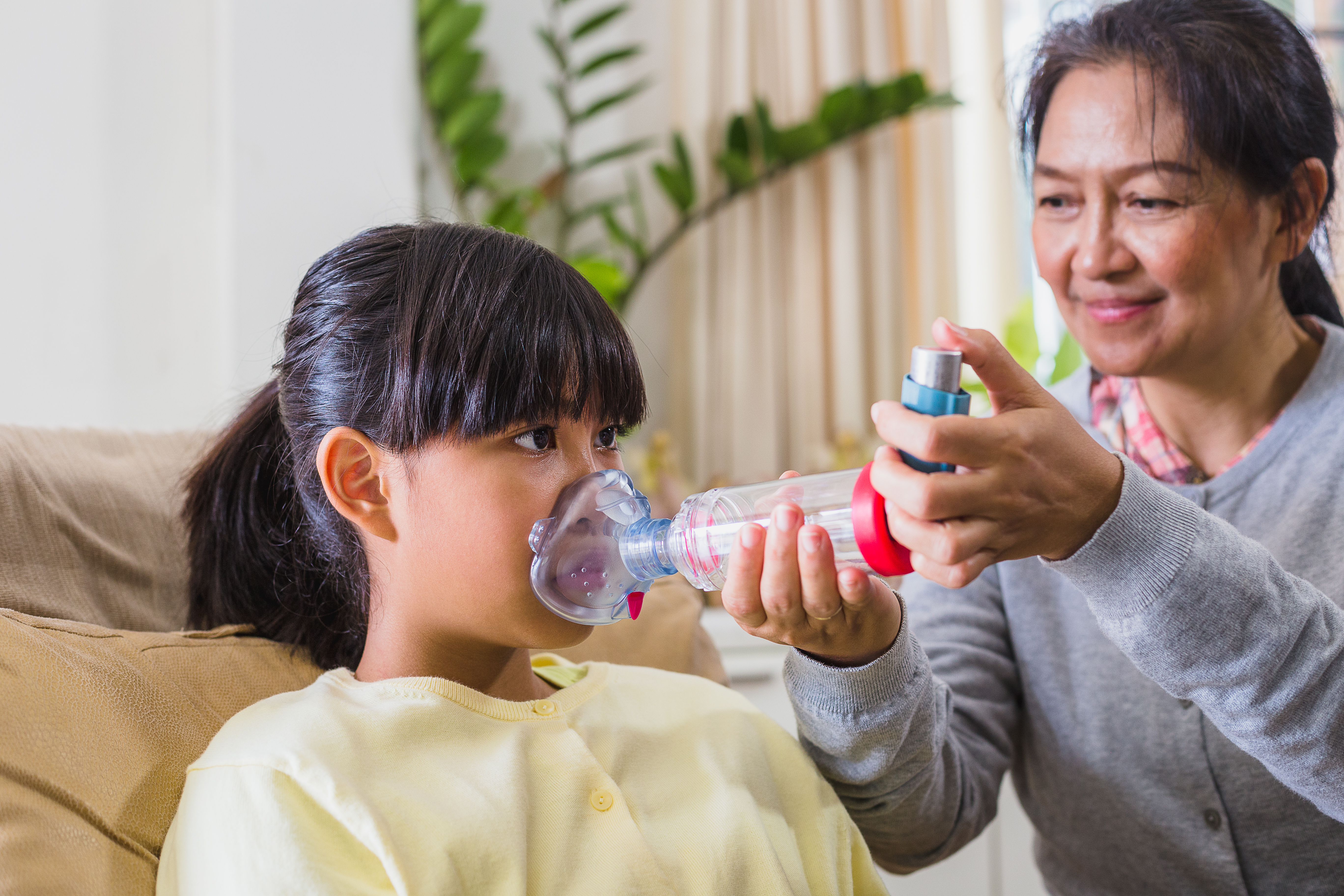 Mother helps her daughter gets assistance with asthma inhaler spacer while setting on sofa in living room at home.