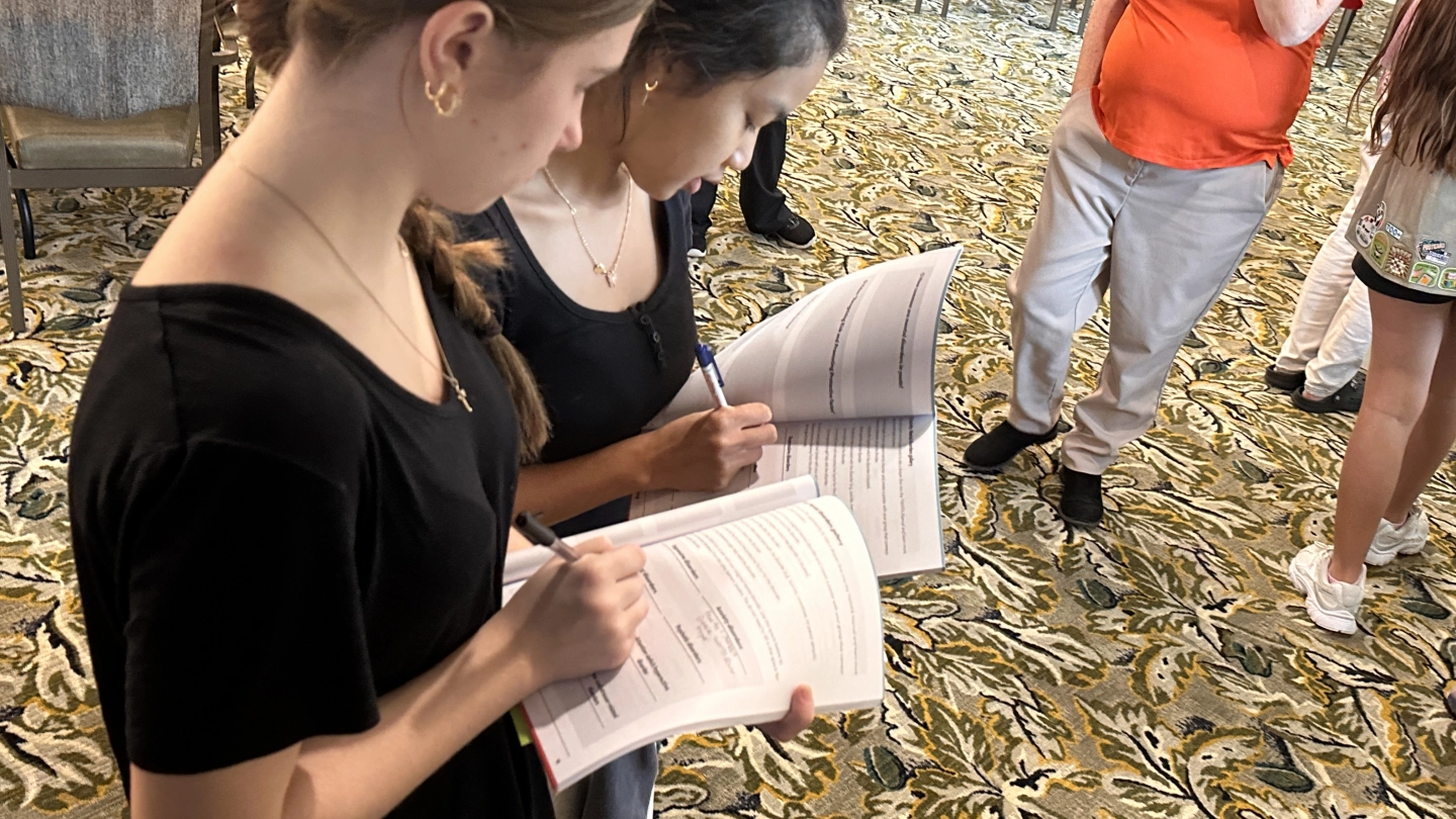 Two individuals stand side by side, writing in booklets during a Youth Mental Health First Aid training session. The indoor setting includes patterned carpet, chairs, tables and other participants engaged in activities.