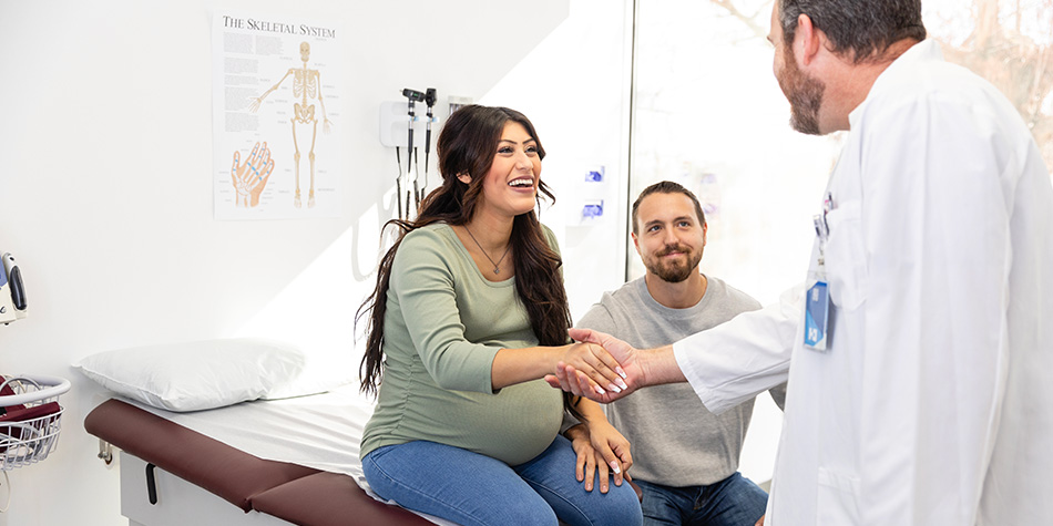 A pregnant woman sitting on an exam room table, shakes her doctor's hand, with her partner sitting next to her.
