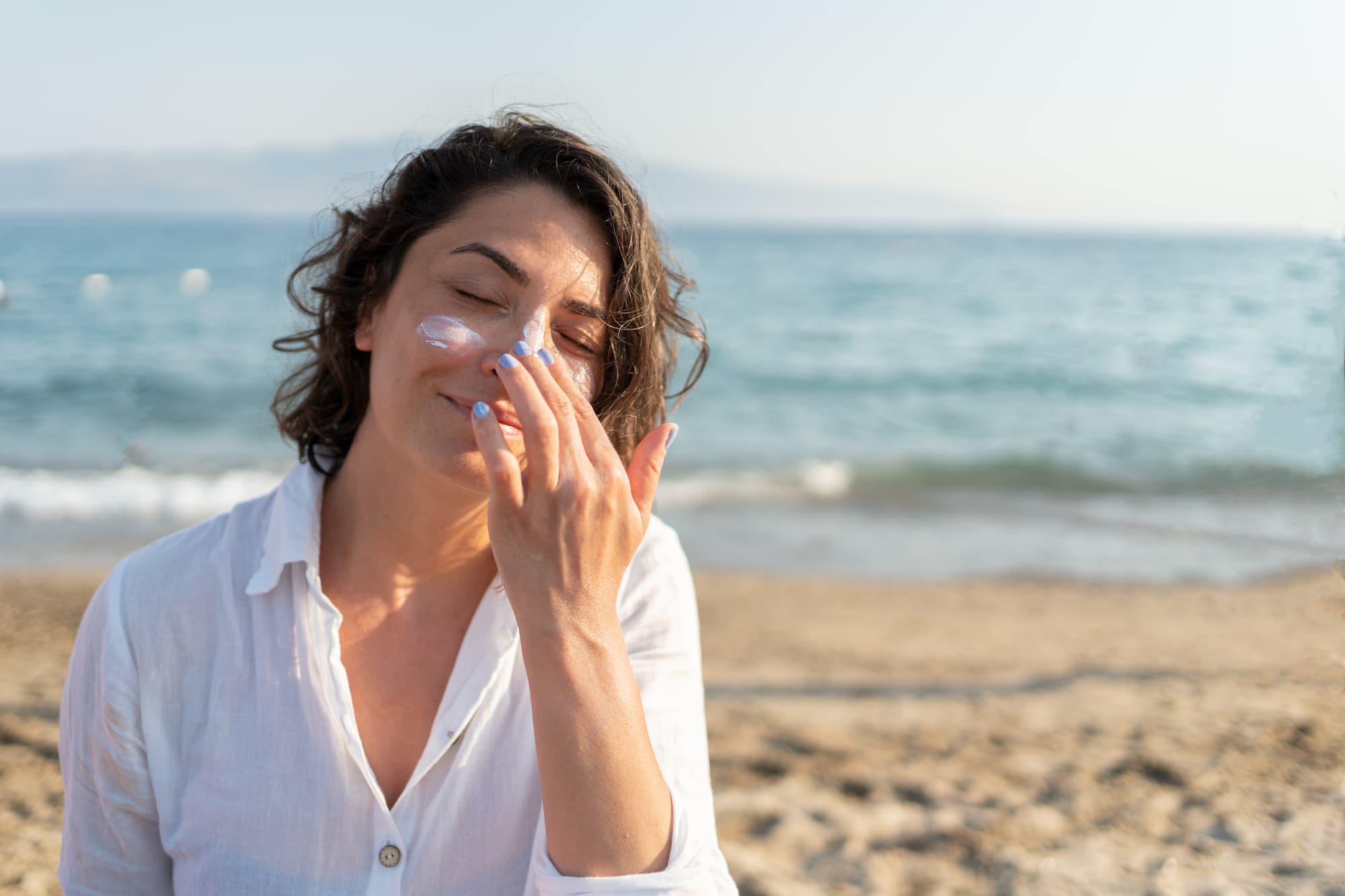 A woman applies liquid sunscreen to her face at the beach.