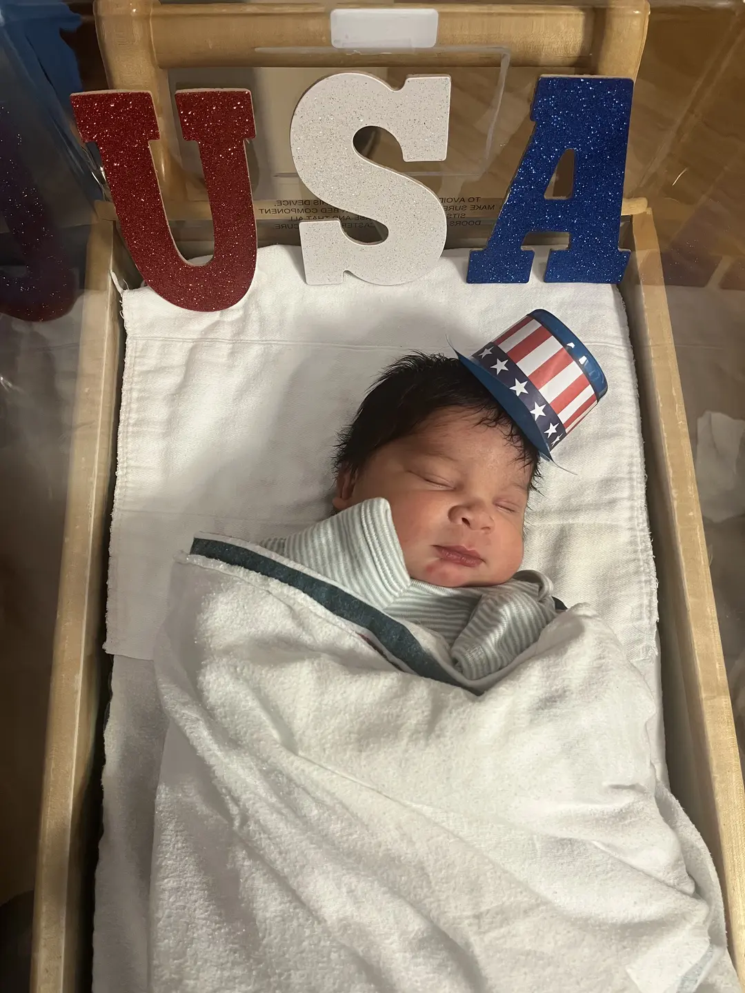 A newborn baby sleeping with a patriotic top hat and the decoration of red white and blue "USA" letters in crib.