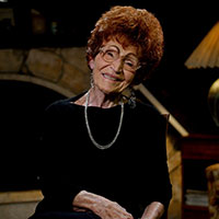 Ramona smiles while sitting in her living room, wearing a black shirt and gold necklace.