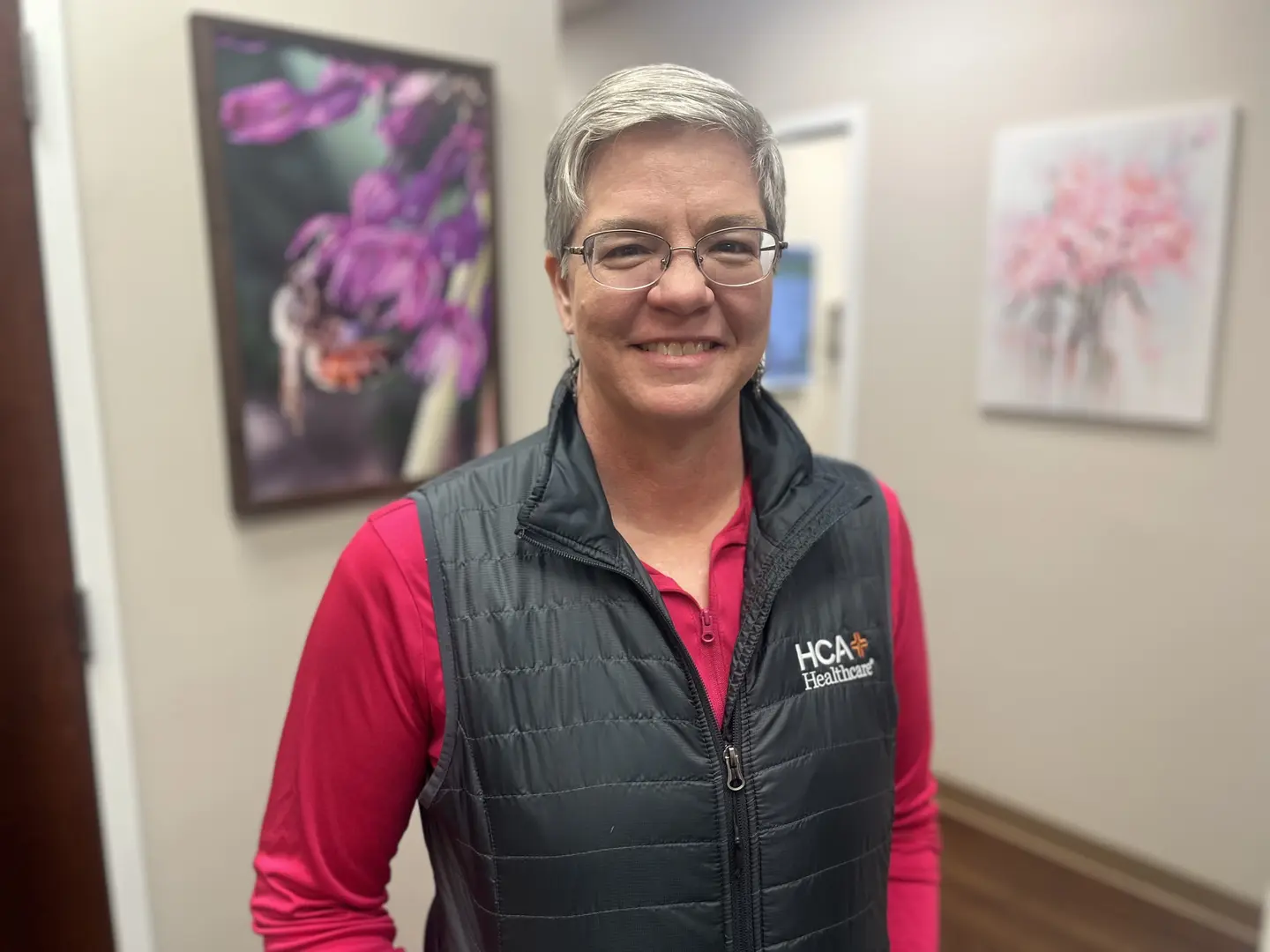 Dr. Jolene Henshaw is standing in a hallway at a LewisGale hospital. She has short blonde hair and is wearing a red shirt with a vest over it.