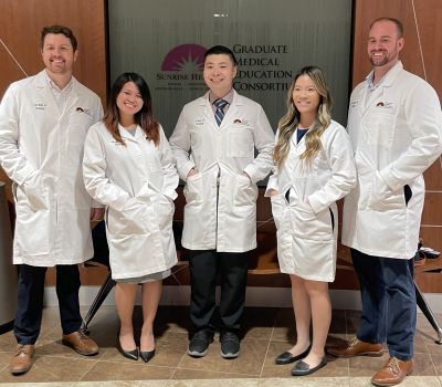 The graduating class, five smiling students in group pose with hands in their white lab coats.