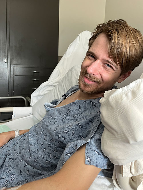 Noah Weaver smiles while lying in a hospital bed, wearing a hospital gown.