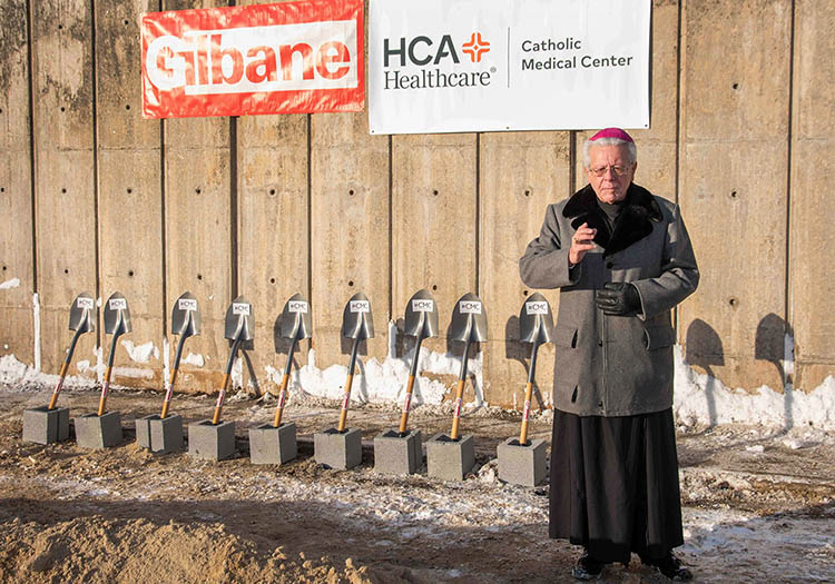 A Catholic Bishop performs a blessing at a groundbreaking ceremony, with CMC-branded shovels resting on cinderblocks in the background.