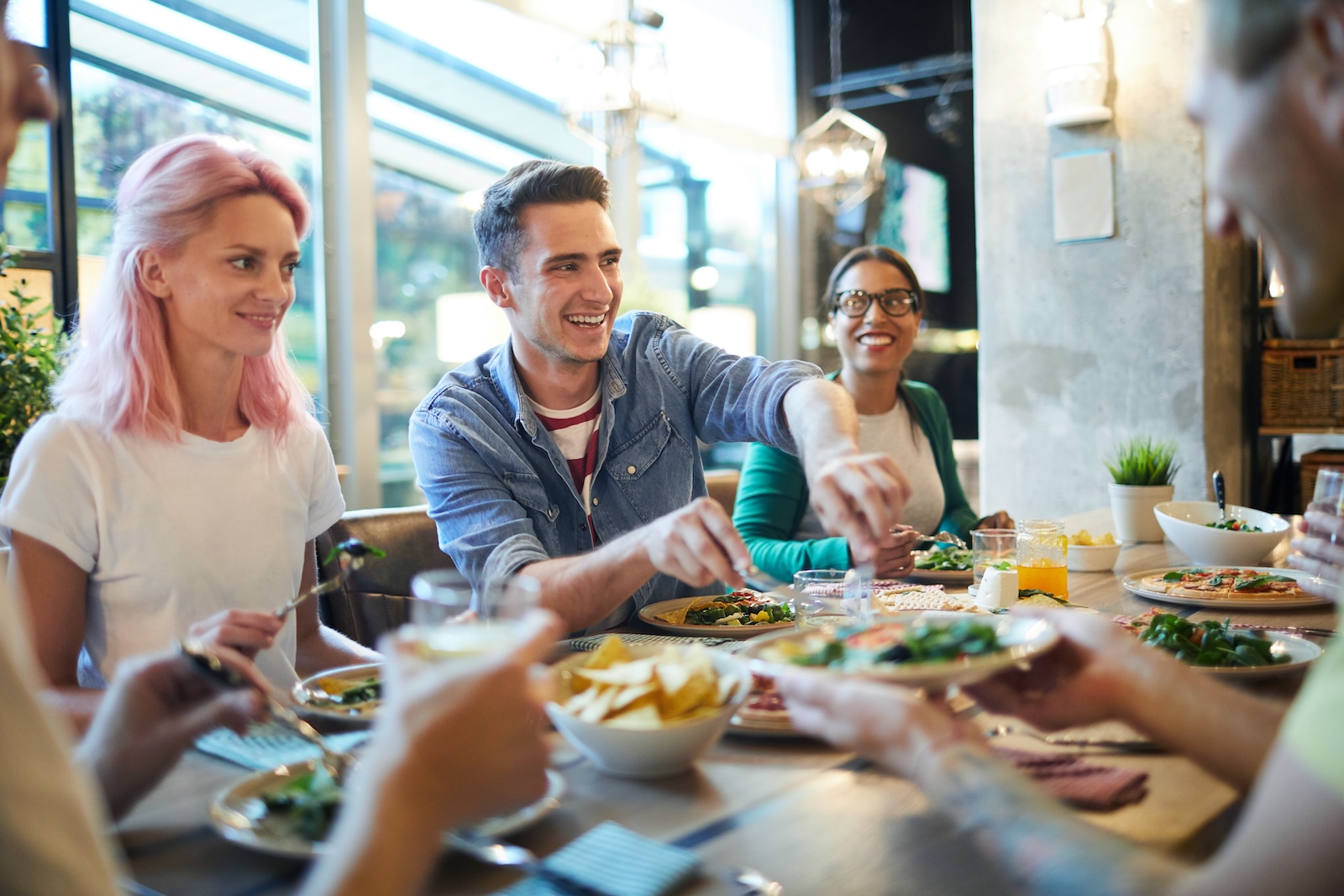 A group of people smiling and laughing at a dinner table, while eating their meals and passing around food to one another.