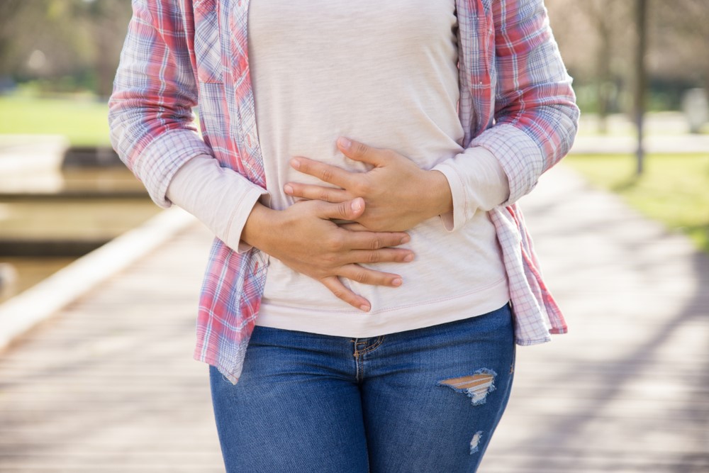 A woman in a plaid shirt, t-shirt and jeans clutching her abdomen outdoors.