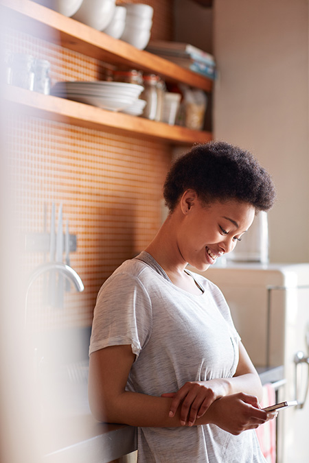 A young woman leans on the kitchen counter by the sink, smiling as she looks at her phone.