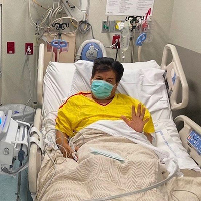 Jose waves to the camera, while lying in a patient bed wearing a medical gown and mask, following his kidney transplant.
