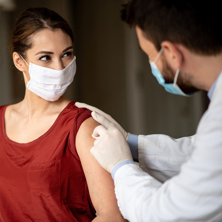 A medical professional is swabbing and preparing a female patient's upper arm for a shot. Both are wearing face masks.