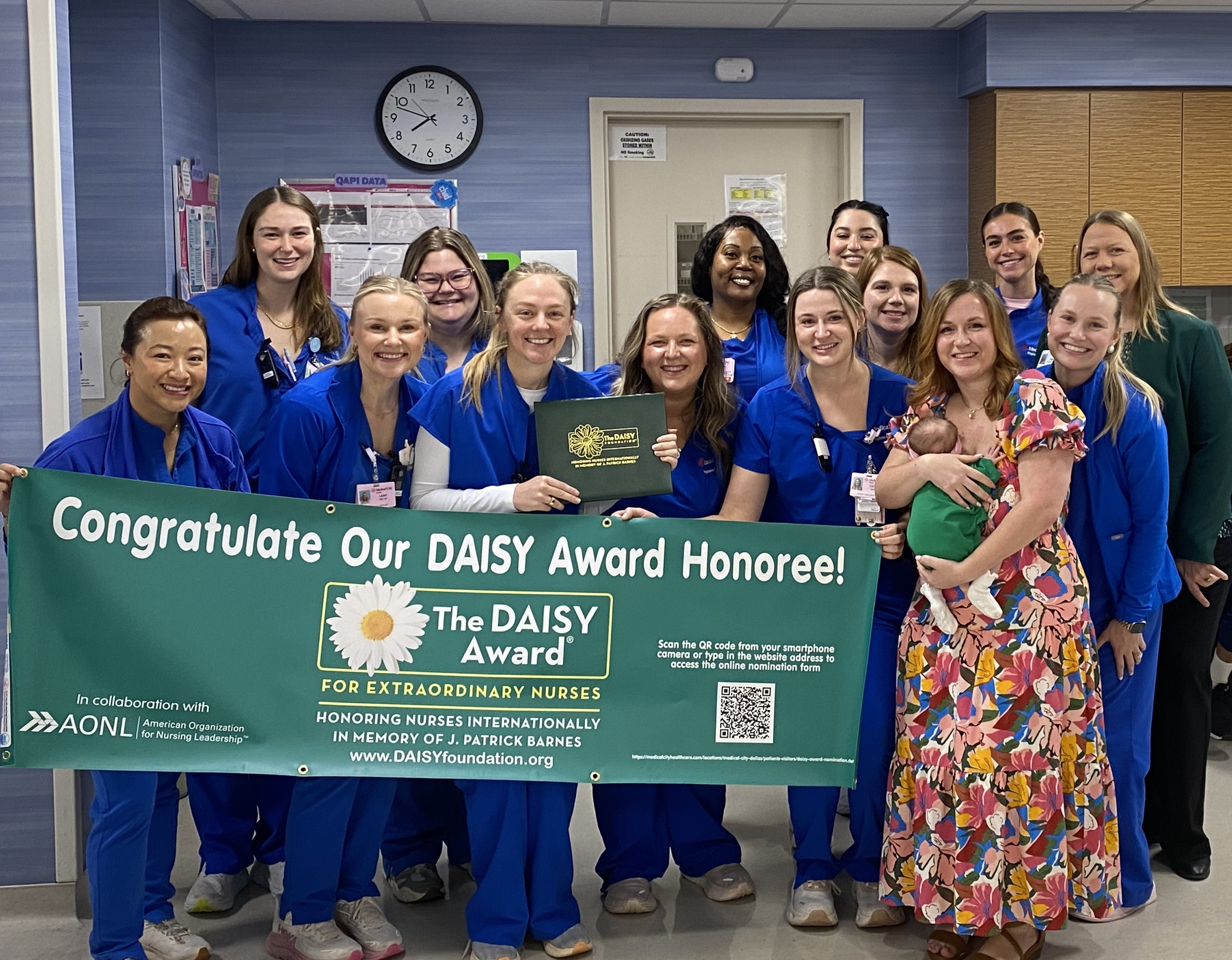 Group of hospital nurses in blue scrubs standing together in a patient care unit, holding a green "DAISY Award for Extraordinary Nurses" banner and certificate. A mother holding her baby stands beside the honored nurse as the team smiles and celebrates the DAISY Award recognition.