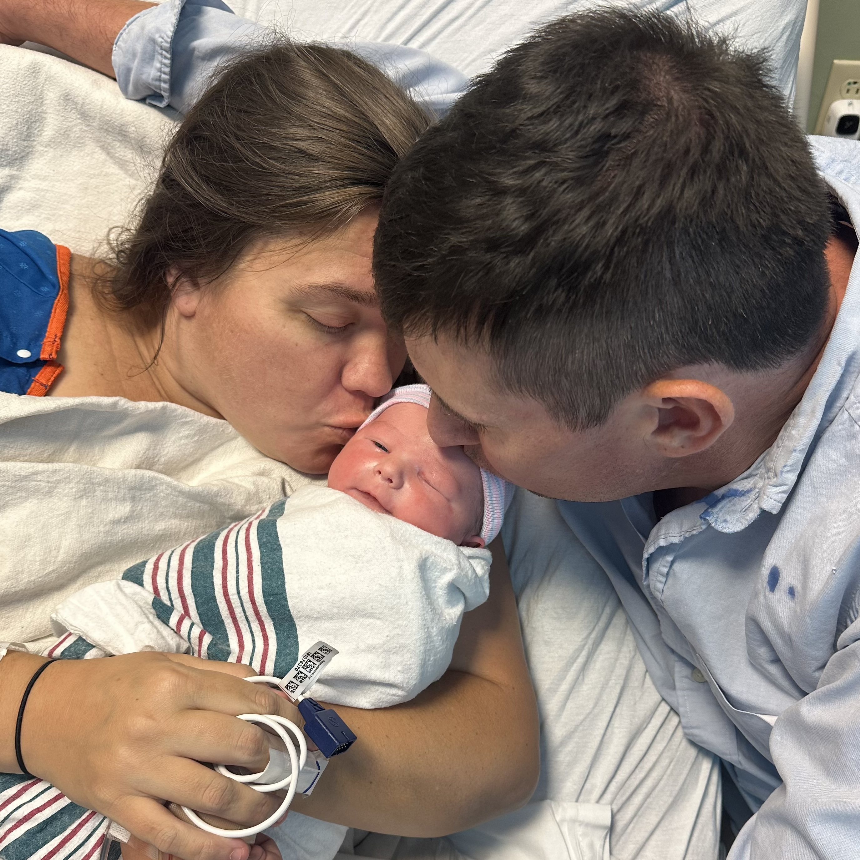 Photo showing of a mom-who just delivered her son, holding him close, her husband next to her; both of them kissing the newborn baby.