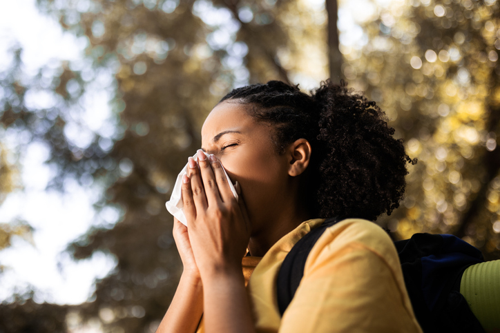Young adult woman with curly hair sneezing into a handkerchief, while standing outdoors.