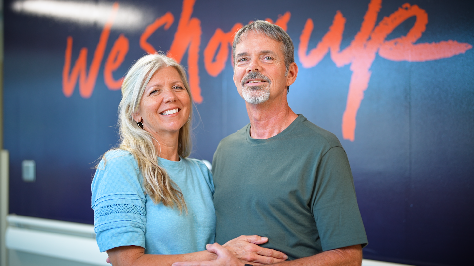 A woman in a light blue shirt and a man in a green shirt embrace in front of a dark blue sign.