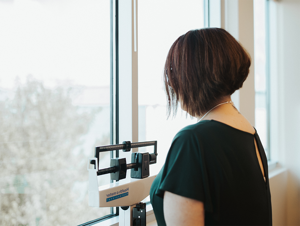 A woman stands on a scale to be weighed, in front of a hospital window.