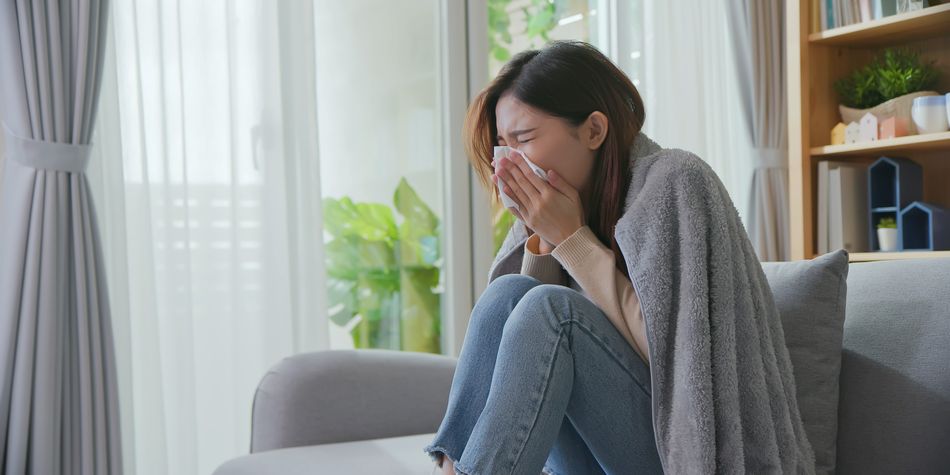 An ill woman is blowing her running nose into a tissue, while seated on a sofa in a living room.