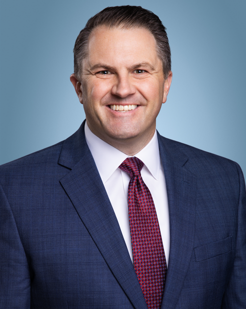 Head-and-shoulders business portrait of Ryan LeMasters, chief executive officer at TriStar StoneCrest Medical Center, smiling and wearing a navy suit, white dress shirt and red patterned tie against a pale blue background.
