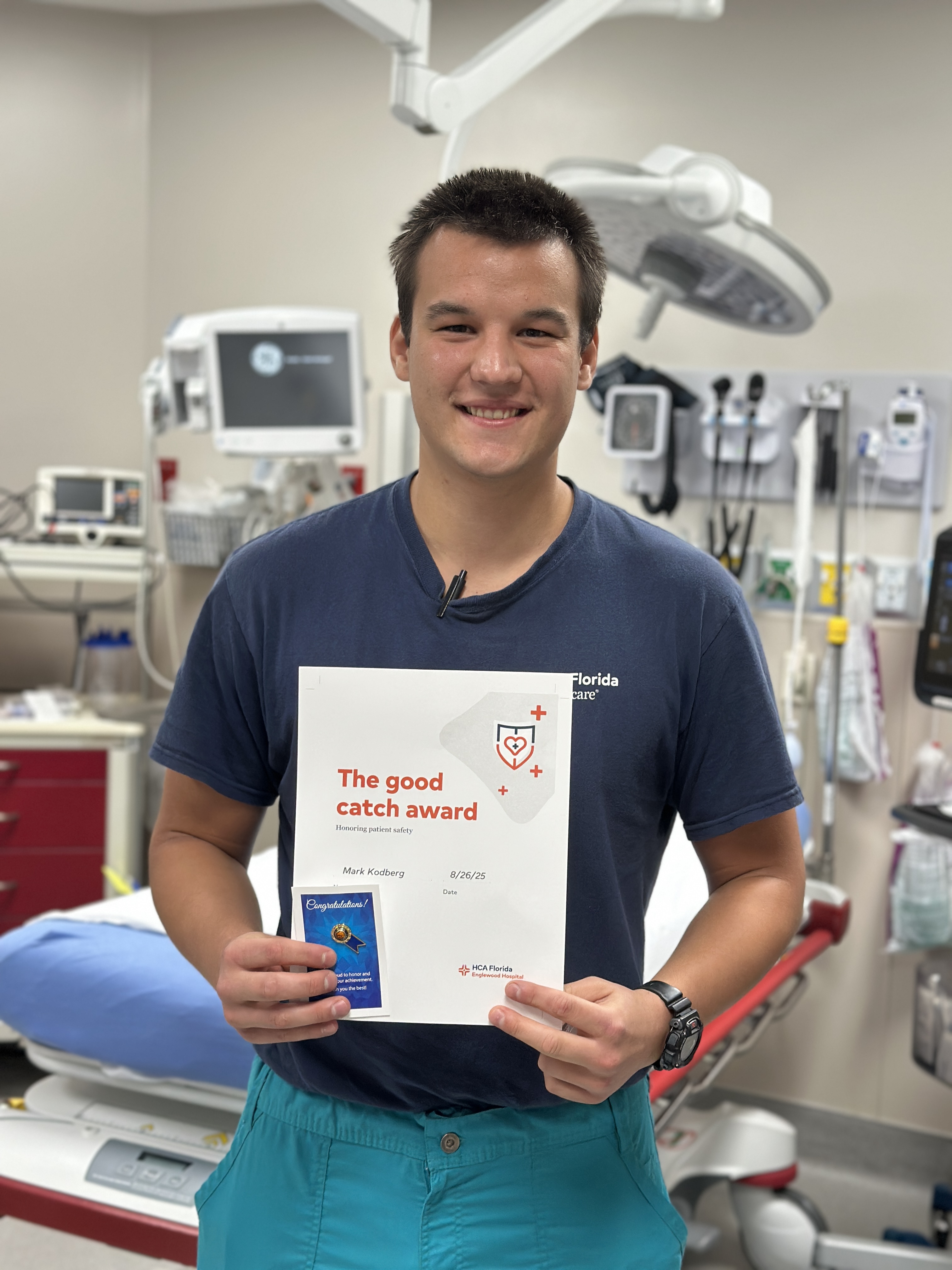 A male nurse stands in a hospital emergency department exam room, holding a certificate recognizing him for identifying stroke symptoms in a patient and helping initiate rapid treatment.
