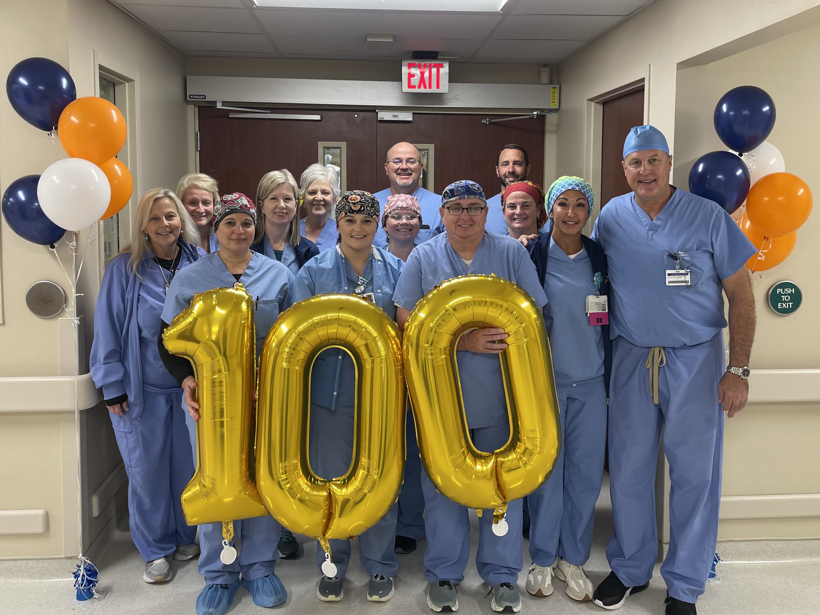 A group of people wearing scrubs smiling and holding balloons that say "100"