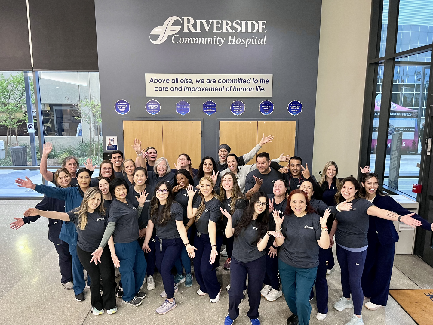 Group photo of Riverside Community Hospital care team members in the hospital lobby beneath the hospital name and mission statement, smiling with arms raised in celebration for G5 Ortho Trauma receiving a 2025 HCA Healthcare Unit of Distinction honorable mention in medical-surgical care.