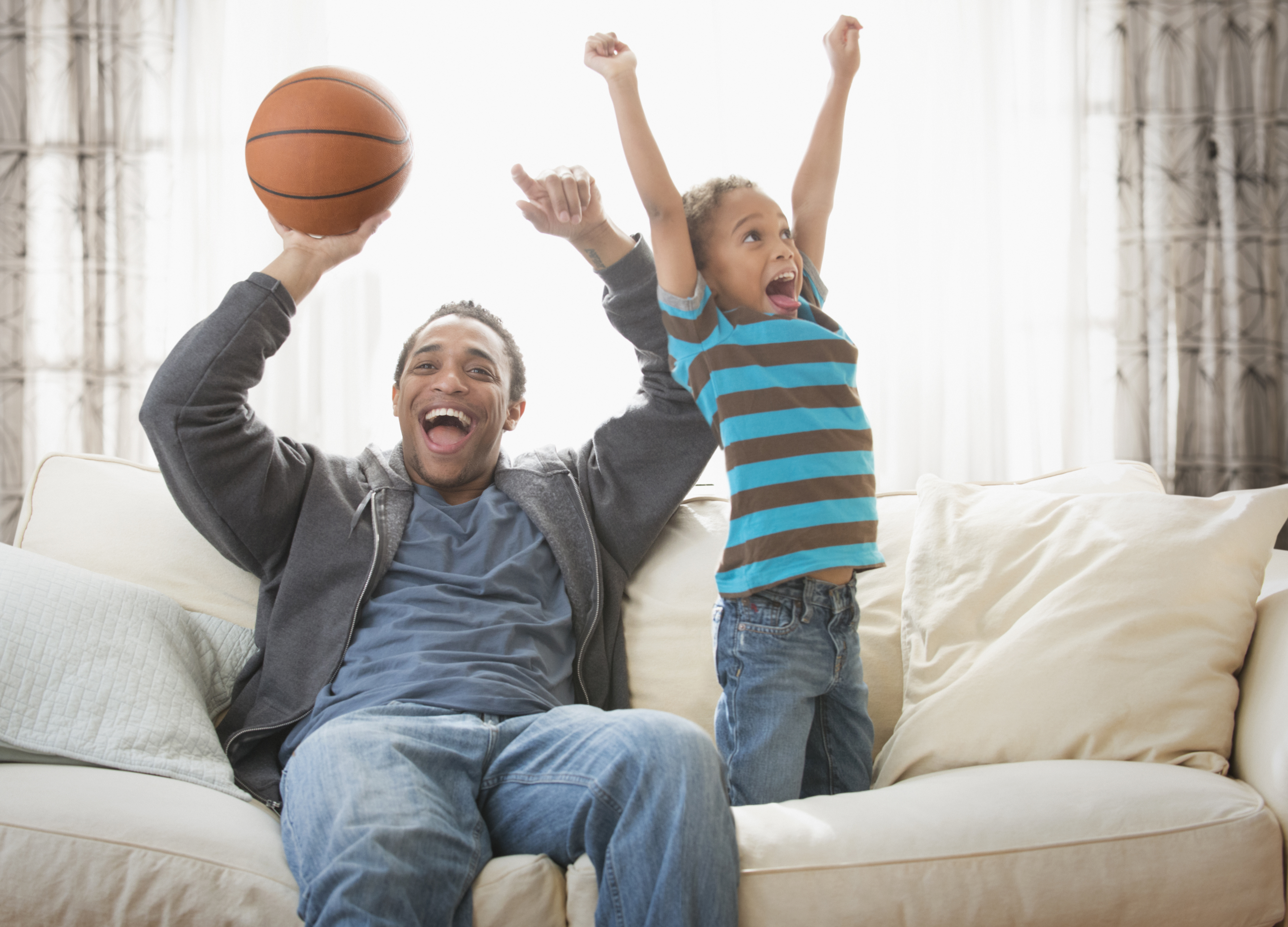 A smiling father holding a basketball and his son raise their arms to cheer, while watching a game from the couch.