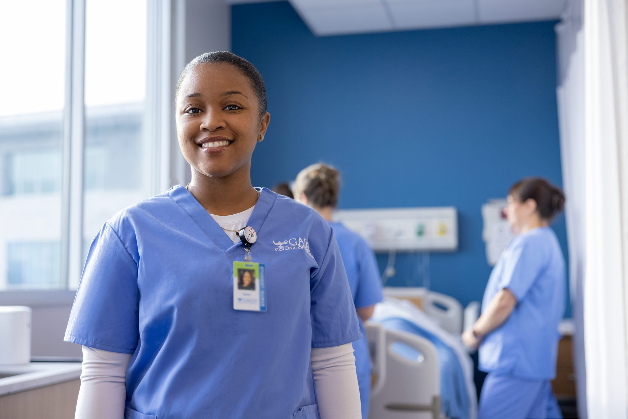 A smiling nursing student in light blue scrubs stands in a bright clinical training room, facing the camera. She wears a student nurse ID badge, while two other healthcare students work near a hospital bed in the softly blurred background. Large windows provide natural light, and a blue accent wall gives the space a clean, professional healthcare setting.