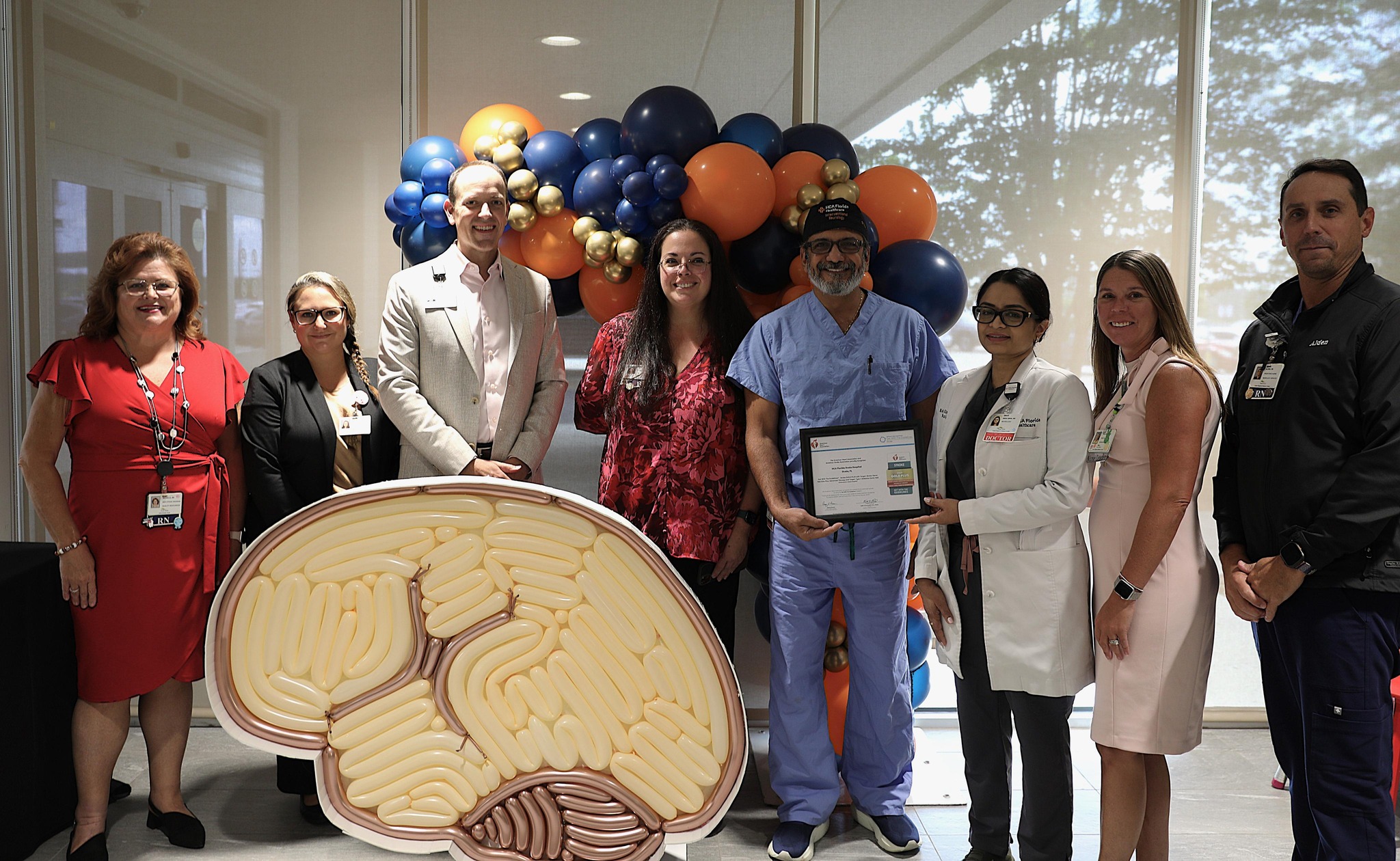 A group of five women and three men stand in front of a colorful display of orange and blue balloons and hold a certificate.