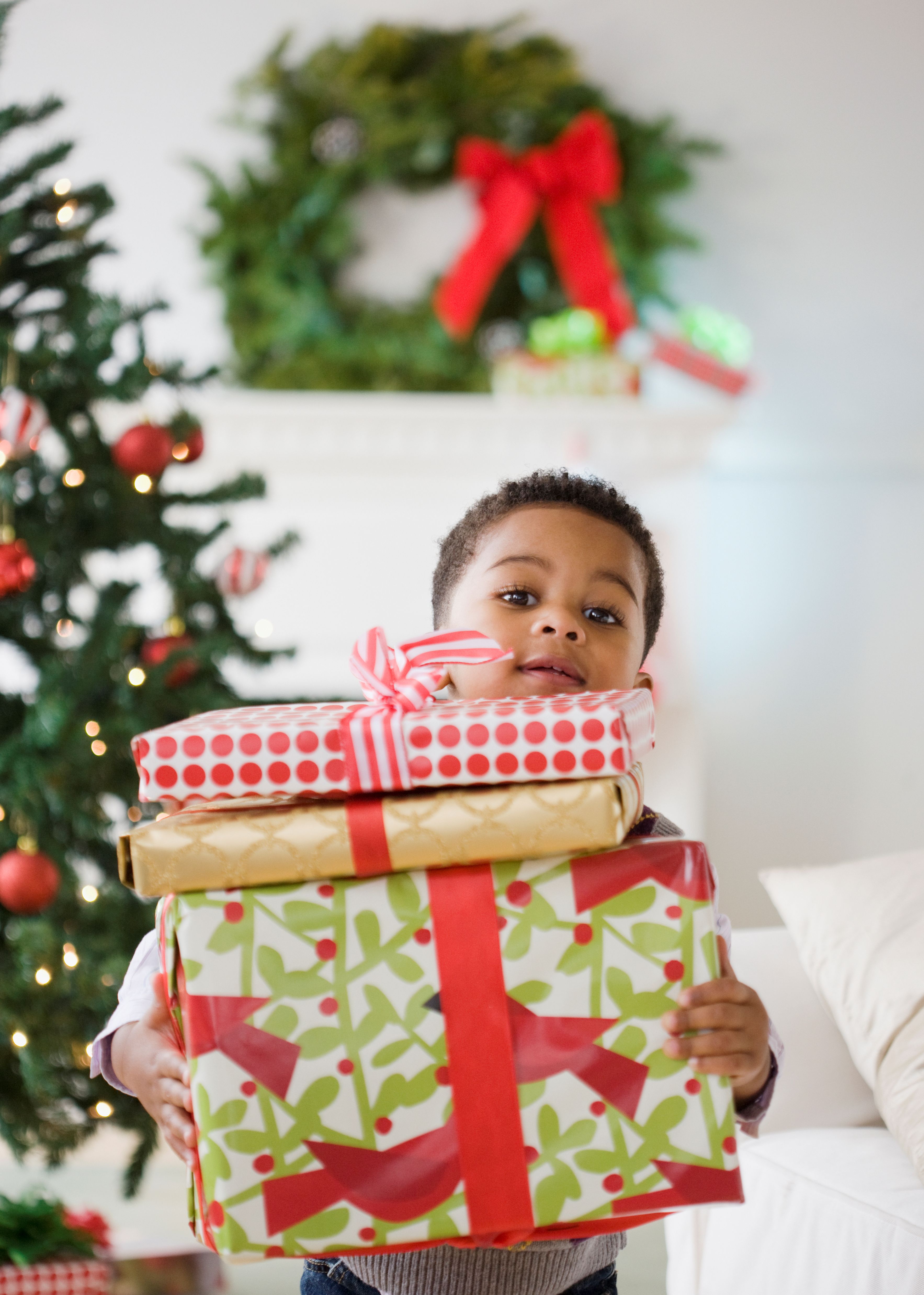A child standing next to a Christmas tree while holding three gifts.