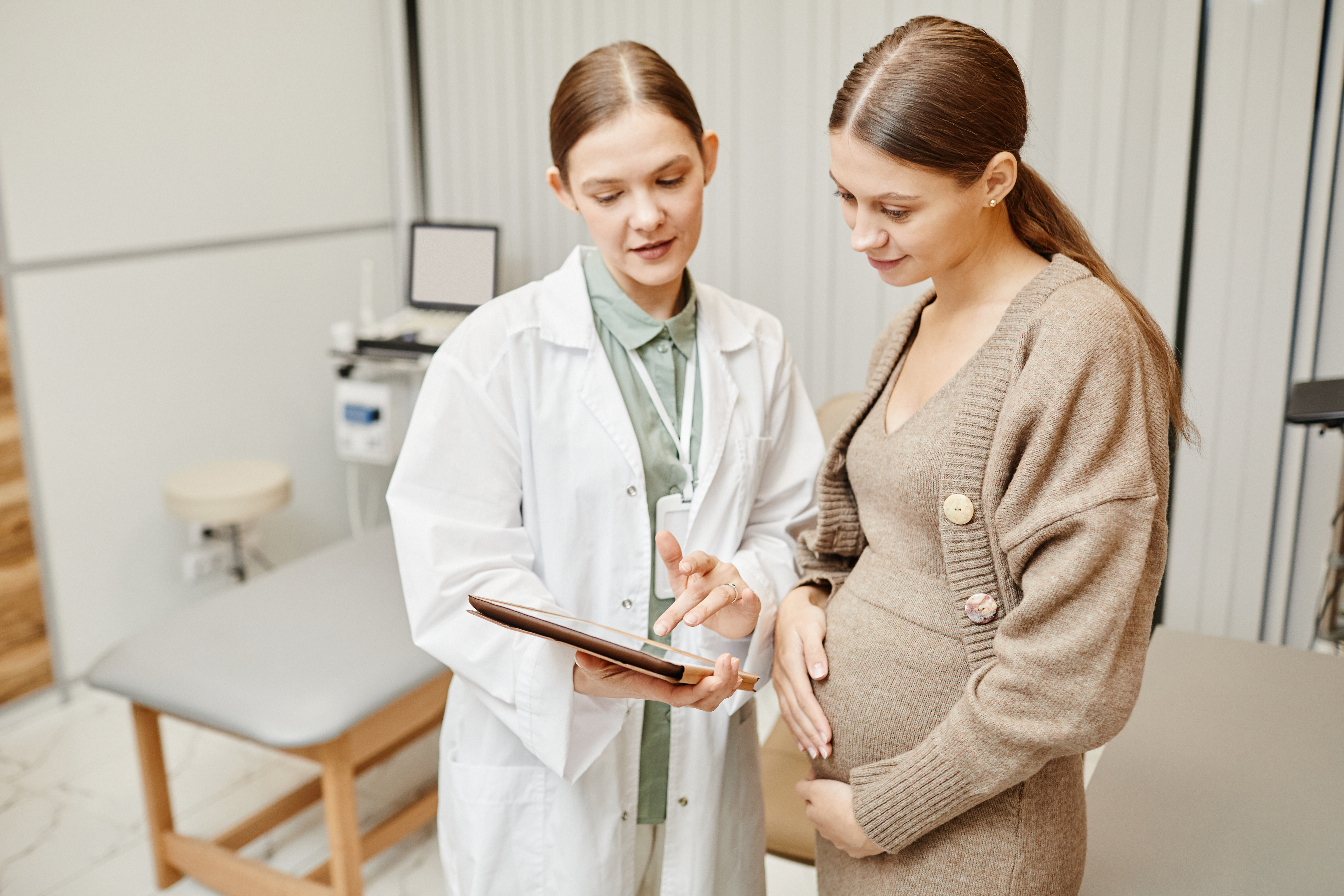 A doctor and a pregnant patient standing while looking at a tablet in a patient room.