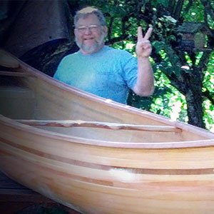 Charles Amspacher holding his arm up, making the peace sign, beside a wooden canoe outdoors. 